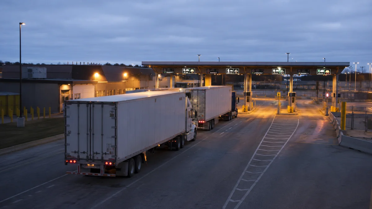 Transport trucks queued in a commercial inspection lane at a Canada-United States border crossing at dusk, with empty inspection booths lit ahead of them.