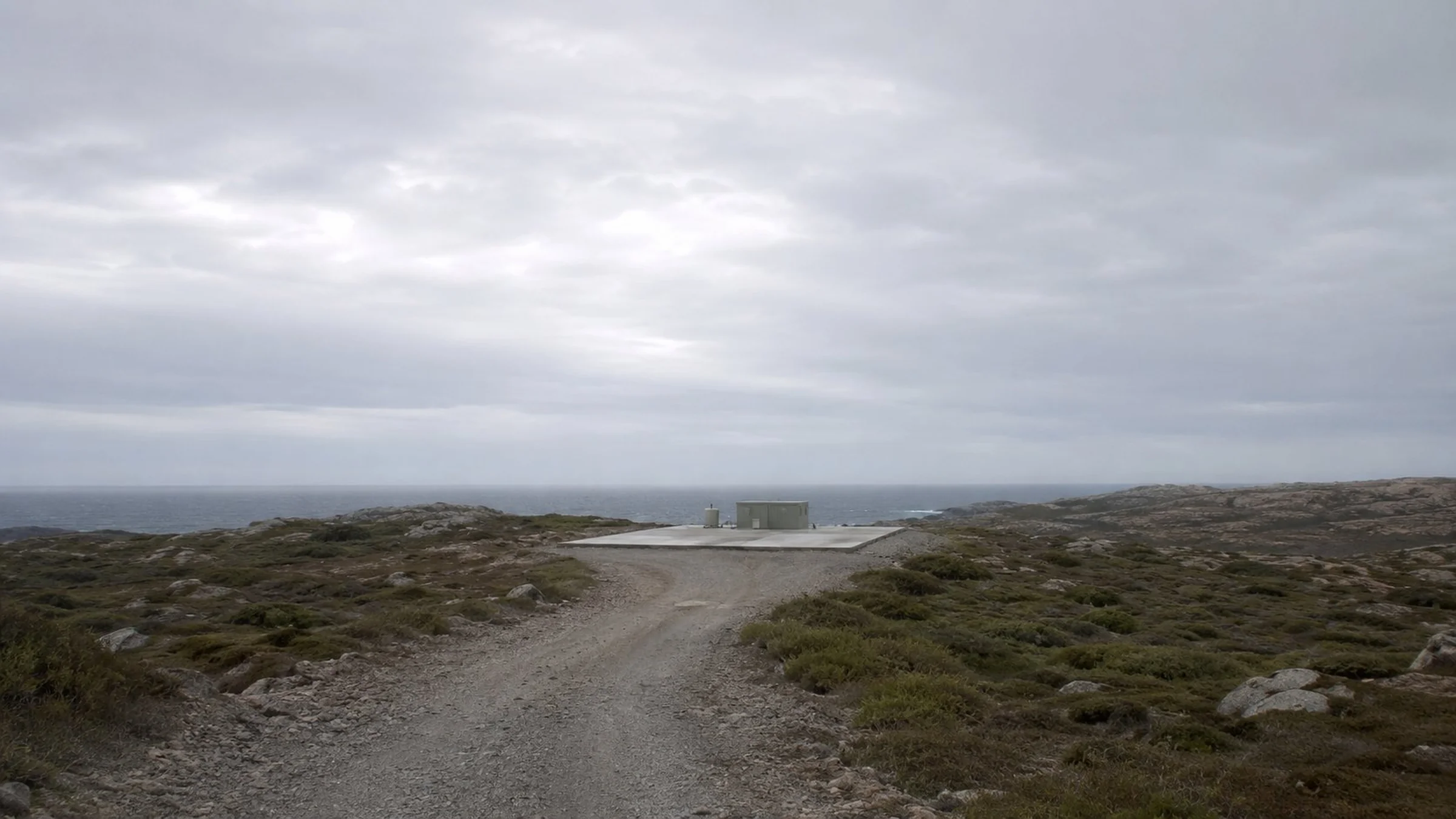 A single concrete launch pad with one low utility structure at the end of a gravel road, on a remote windswept Atlantic coast under a large overcast sky.